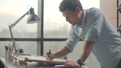 Un-Hombre-Es-Un-Joven-Arquitecto-Moderno-Sentado-En-Una-Mesa-Dibujando-Un-Plano-Del-Edificio-Y-Desarrollando-Un-Diseño-Sentado-En-Una-Oficina-En-El-Loft-Del-Sitio-Cerca-De-Una-Gran-Ventana.-Crear-Fachadas-De-Edificios,-Diseñar-Muebles-Y-Diseñar-El-Proyecto-De-Un-Conjunto-Residencial.