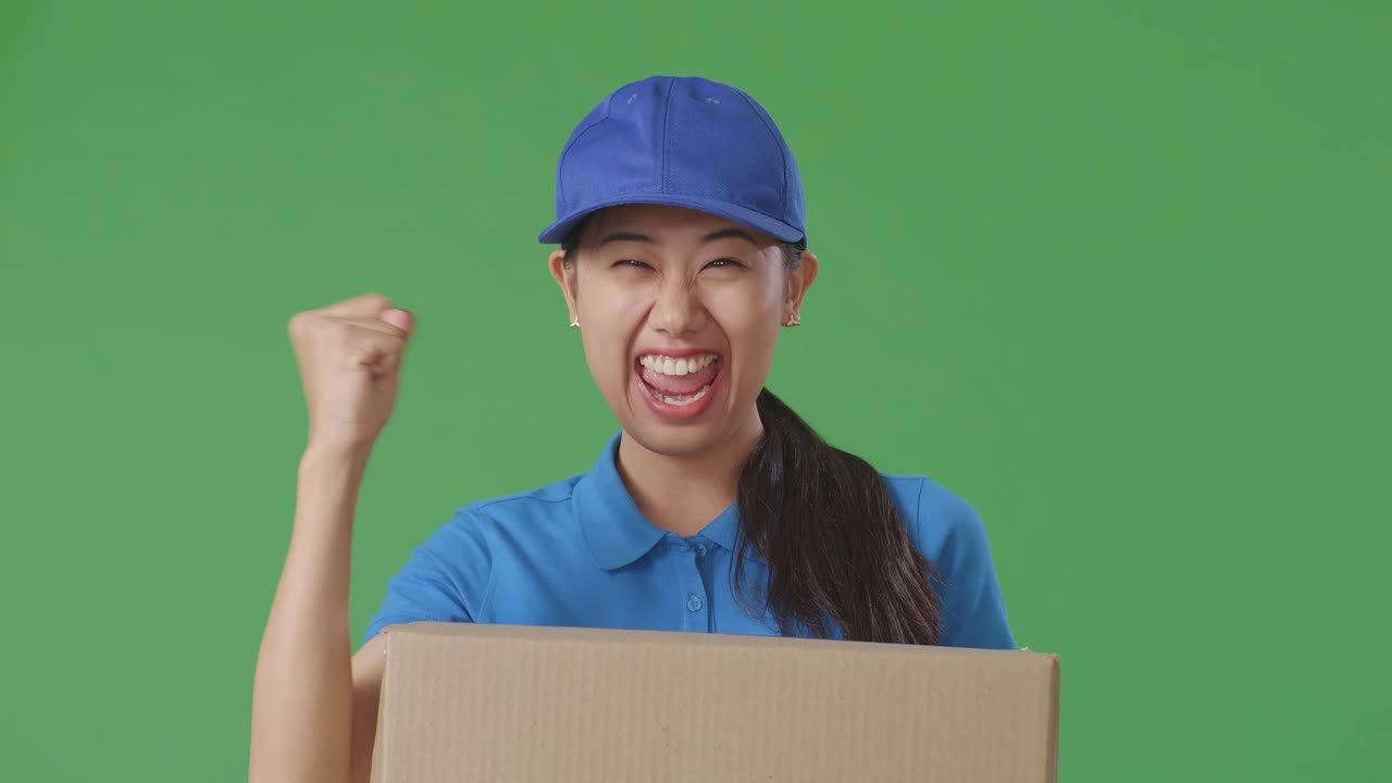 푸른 유니폼을 입은 아시아 여성 택배사가 성공을 축하하며 스튜디오의 녹색 스크린 배경에 박스를 배달합니다.