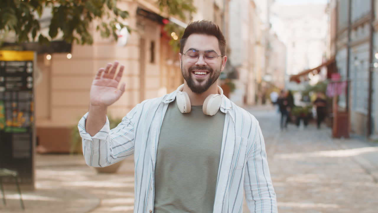 joven alegre sonriendo amistosamente a la cámara agitando las manos gestando mientras está de pie en la calle de la ciudad