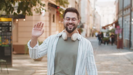 cheerful young man smiling friendly at camera waving hands gesturing while standing on city street