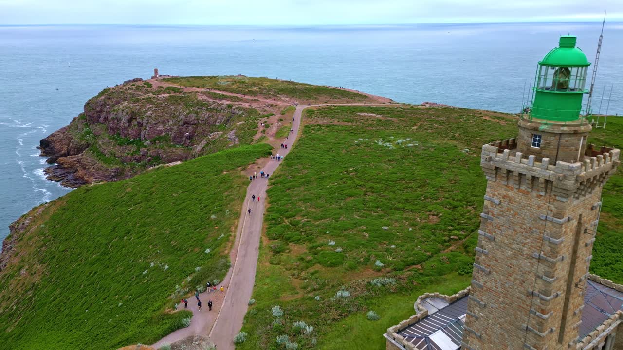 Historic Cap Fréhel lighthouse and old Vauban tower on rugged cliffs, people walking path, Brittany, France, Scenic coastline, travel. Aerial forward
