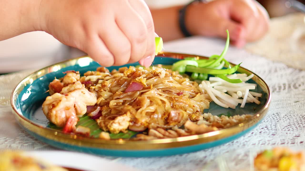 A hand squeezes lime over a vibrant Pad Thai dish, enhancing flavors. Bright lighting highlights the colorful ingredients on a decorative plate