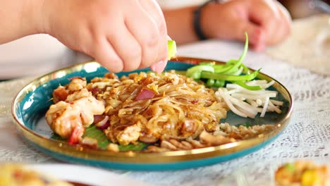 a hand squeezes lime over a vibrant pad thai dish, enhancing flavors. bright lighting highlights the colorful ingredients on a decorative plate