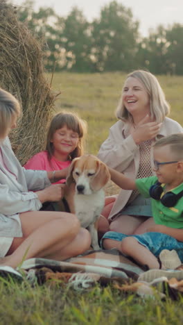 las hermanas con hijos disfrutan de tocar al perro en el campo abierto, sentarse junto a la bala de heno durante la puesta de sol, un cálido momento familiar en un entorno rural, ocio casual al aire libre