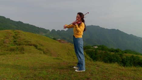 Mujer-Tocando-El-Violín-En-Una-Colina-En-Un-Clima-Nublado-Con-Fondo-De-Montaña