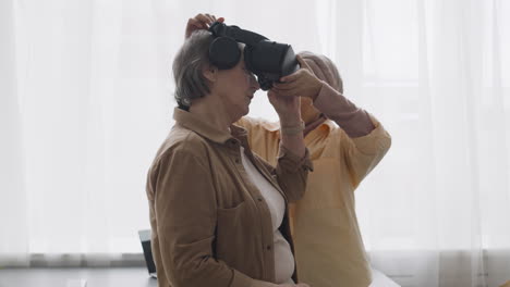 middle aged arabic woman helping a senior lady to use virtual reality headset glasses at home 3