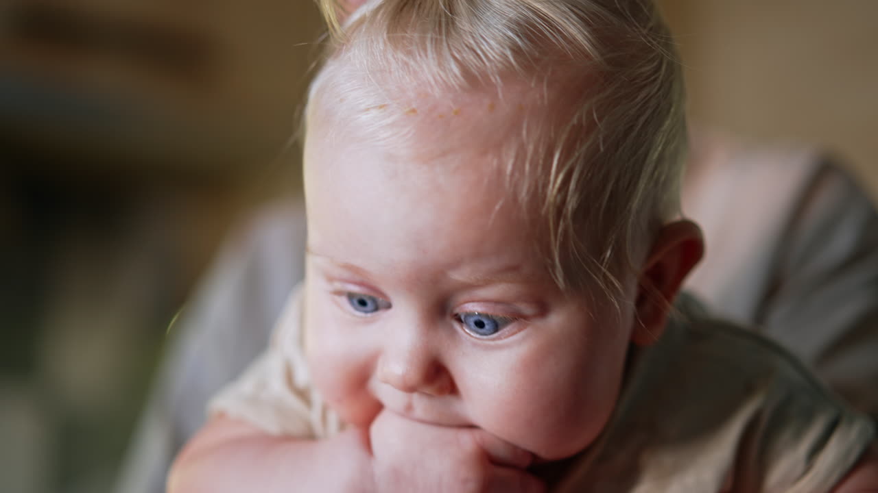 Face of a cute Caucasian blond baby chewing his hand. Close up portrait of an infant child in mom's hands. Blurred backdrop.