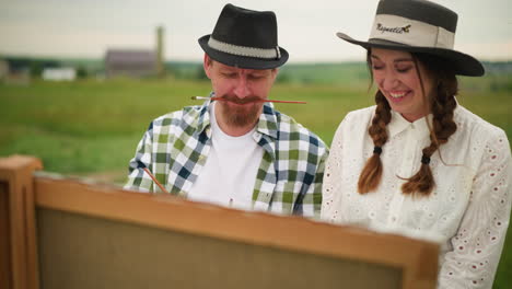 a creative scene featuring a painter wearing a checkered shirt, holding a paintbrush in his mouth while focusing on his work. beside him, a woman in a white dress and hat stands, smiling warmly