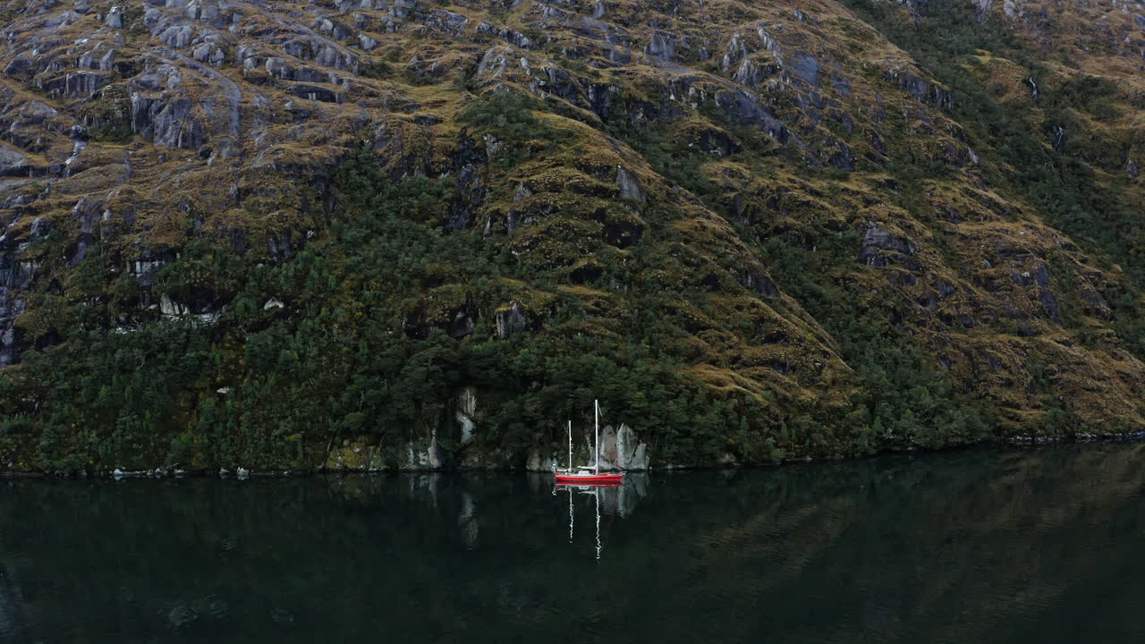 Sailboat nears forested shoreline of Beagle Channel under fading daylight and calm waters, aerial pullback
