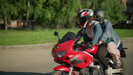 side view of two women riding a power bike through an urban road, the passenger holds onto the rider as they pass by trees and parked cars, with buildings visible through the trees