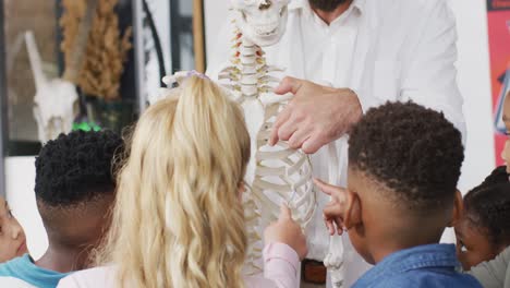 Diverse-male-teacher-and-happy-schoolchildren-studying-skeleton-in-biology-class