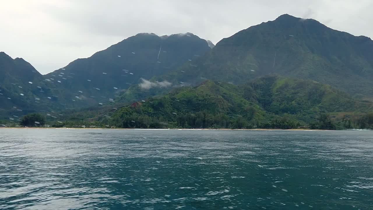 HD 120fps Hawaii Kauai Boating on the ocean floating right to left green hills and mountain with waterfall in cloudy distance with boat spray in foreground