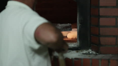 panadero masculino con cáscara de metal deslizando masa de pan dentro del horno de ladrillo tradicional en la panadería