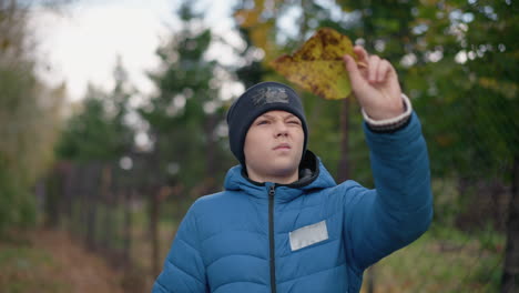 boy in black beanie and blue puffer jacket thoughtfully inspecting dry leaf in hand, standing in front of greenery, drops it, with blurred bar fence in background