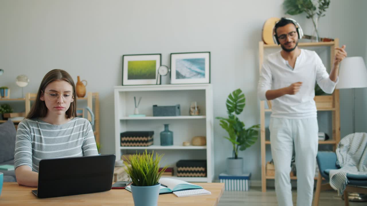 серьезная женщина работает с ноутбуком за столом, в то время как муж танцует в наушниках.