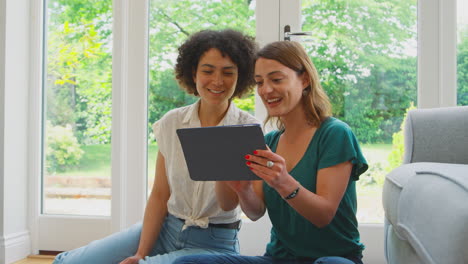same sex female couple or friends at home sitting in lounge using digital tablet
