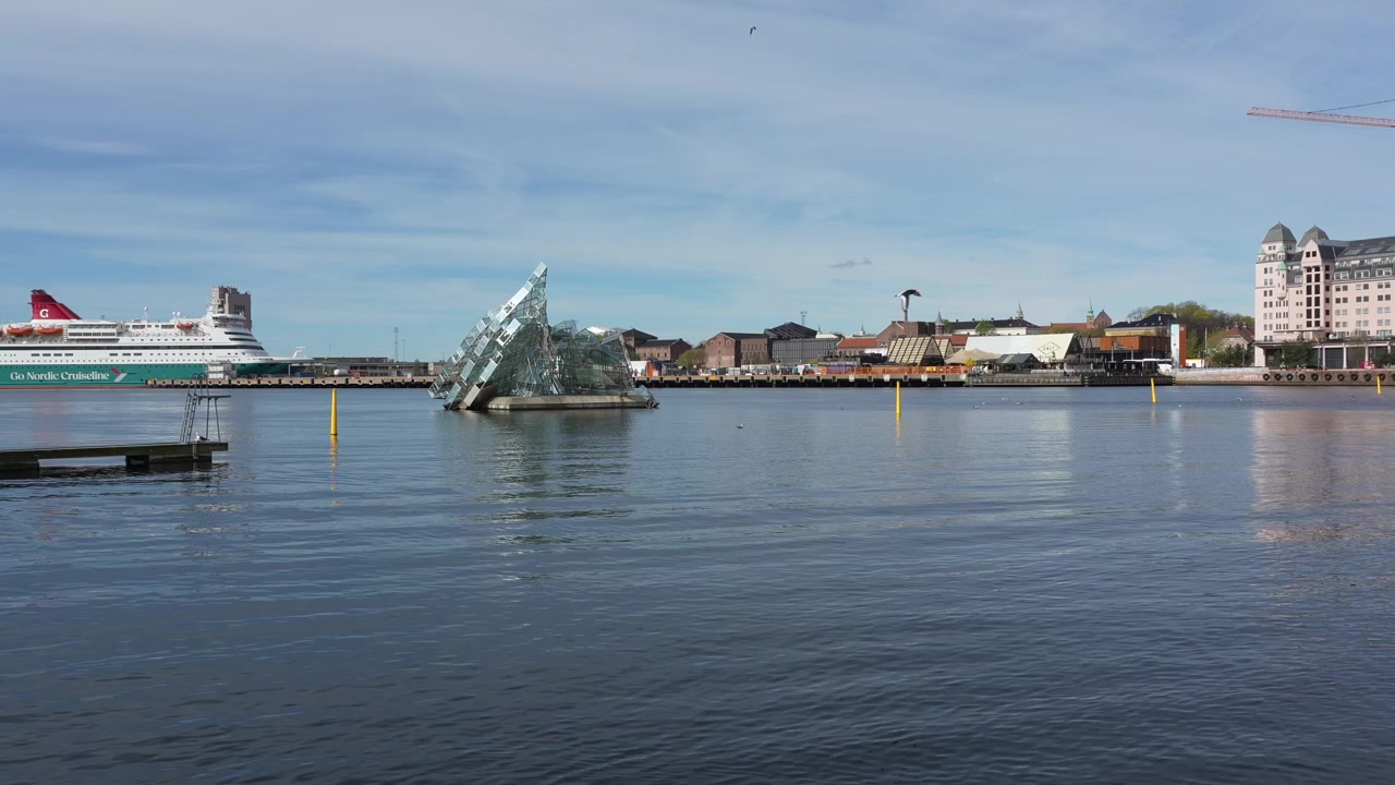 Dzienne ujęcie z ręki zatoki portowej Oslo, przedstawiające rzeźbę „She Lies” na wodzie, nowoczesne bryły Opery, panoramę miasta, nabrzeże, spokojne fale i czyste błękitne niebo.