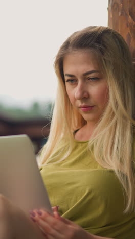 pretty blonde lady tourist watches video via modern laptop computer perching on log handrail of restaurant platform at eco resort on nasty day slow motion