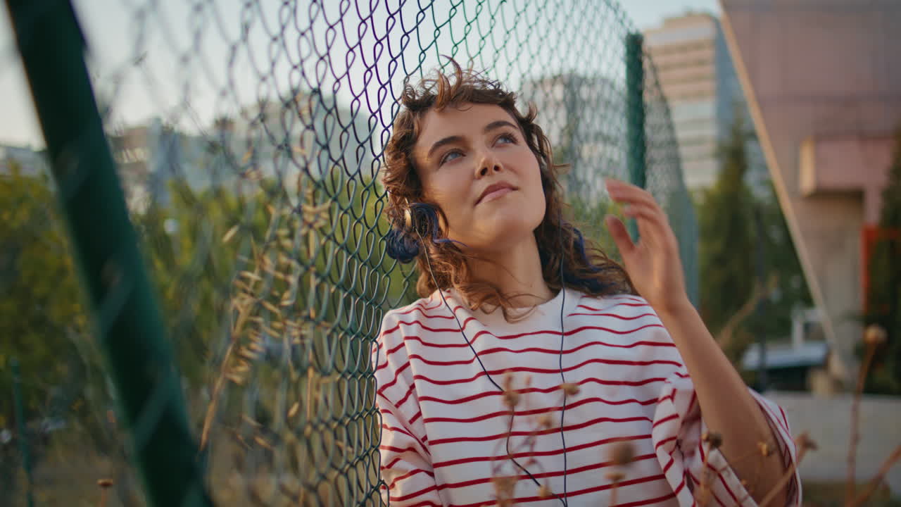 modelo romántico escuchando música inclinándose cerca de malla de metal noche de verano de cerca.