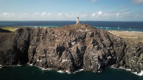 un faro situado en acantilados rocosos con el océano y la aldea distante en el fondo, vista aérea