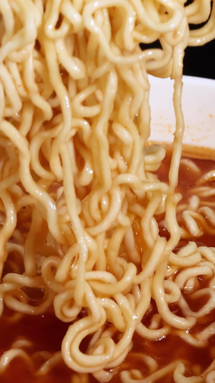 Bacchette afferrano e sollevano noodles istantanei cotti da una ciotola di brodo rosso e piccante sotto un'illuminazione brillante e uniforme con una vista ravvicinata e statica della telecamera