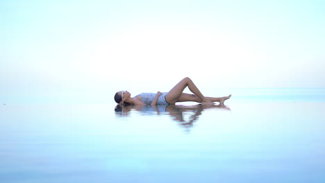 while giving the illusion of floating in mid-air, a fit young woman lies in the shallow edge of an infinity-edge pool