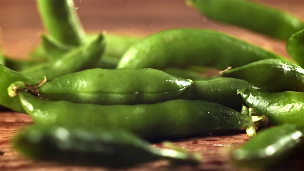 Fresh Green Pea Pods Falling onto a Wet Wooden Surface in Slow Motion