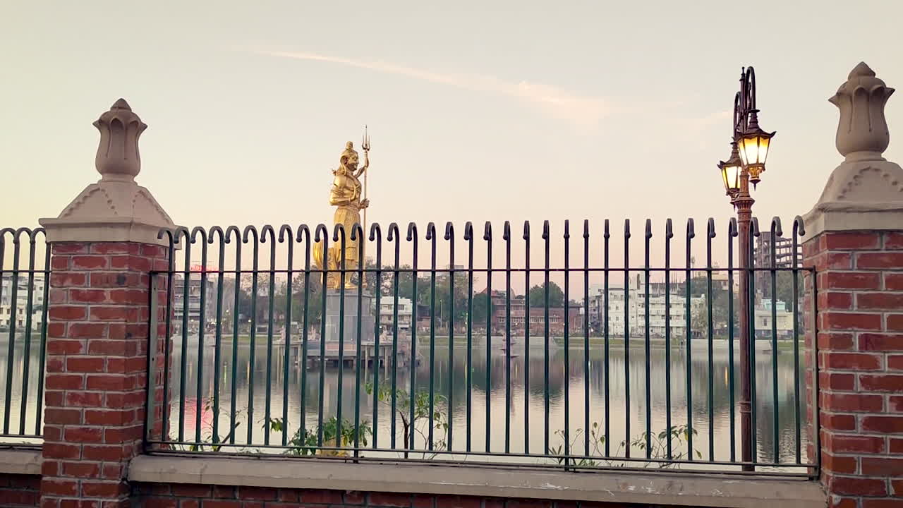 Lago Sursagar, Vadodara, Gujarat, India. Statua dorata di Shiva oltre il lago con recinzione in primo piano e sfondo cittadino, atmosfera diurna calma e punto di riferimento culturale