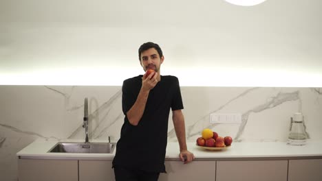 young man eating red apple at home in the kitchen. portrait of guy in black clothes eating a fresh apple and looking at camera. leaning on a counter at modern, white kitchen