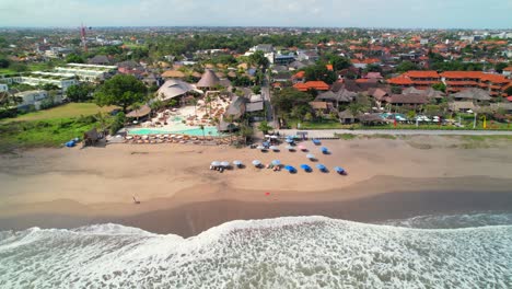 Mari-Beach-Club-Bali-in-Batu-Belig-Beach,-North-Kuta---Aerial-Pull-Back-with-Cityscape-Revealing