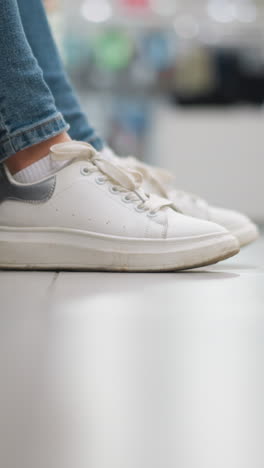 primer plano de la pierna de la dama en vaqueros con zapatos blancos de lona tocando suavemente la pierna derecha en el suelo en un centro comercial bien iluminado, el calzado y el movimiento de las piernas enfatizados en una atmósfera casual y relajada del centro comercial