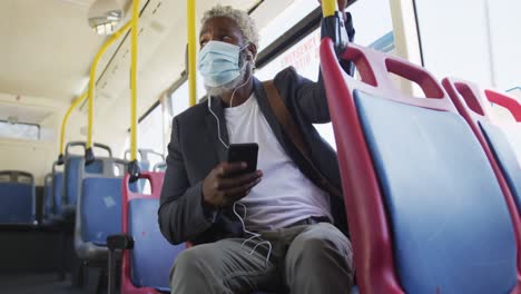 hombre mayor afroamericano con máscara facial usando un teléfono inteligente mientras está sentado en el autobús