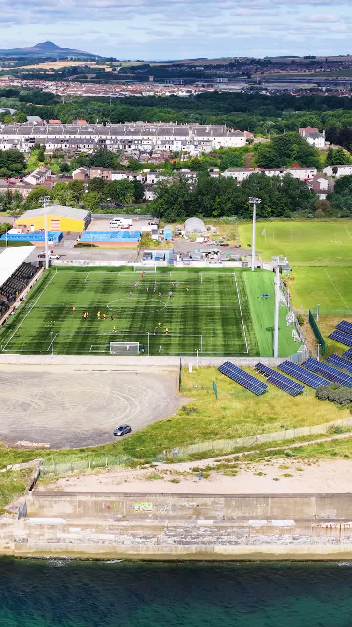Panoramica di un drone su uno stadio di calcio con pannelli solari, campi di allenamento e paesaggio urbano di giorno.