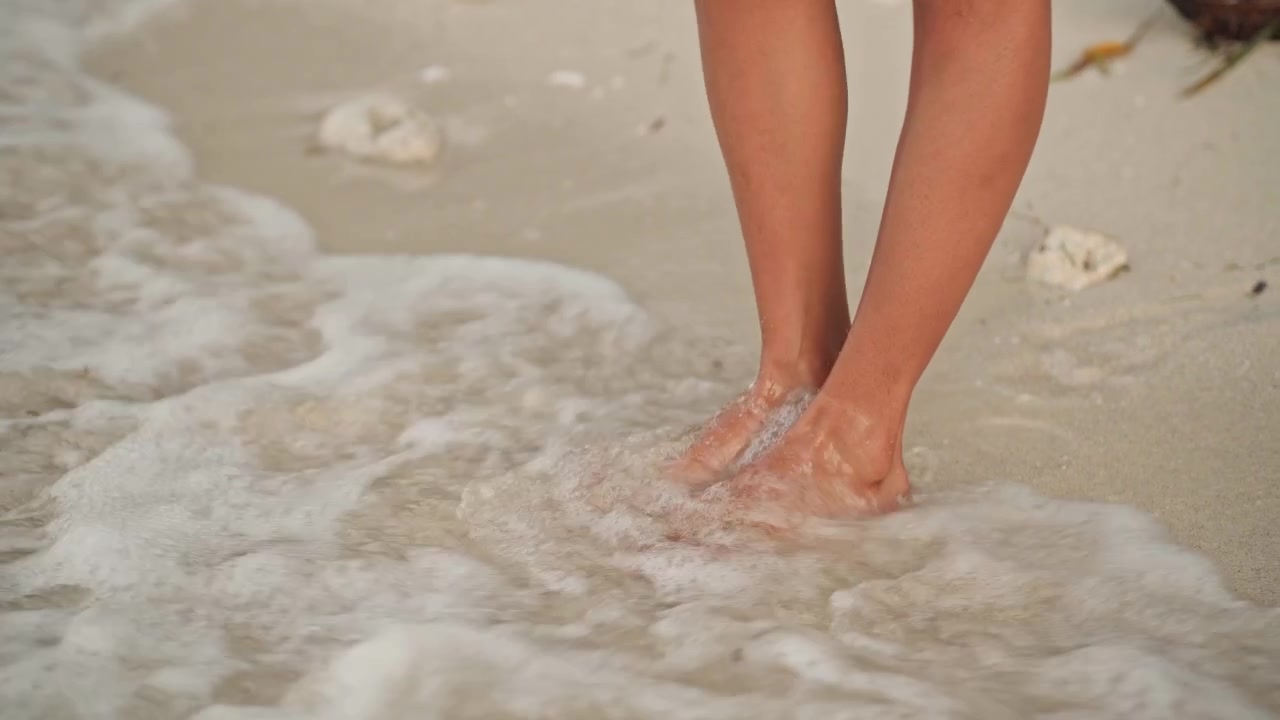 Piedi sulla battigia sabbiosa, onde delicate che lambiscono le dita dei piedi, una vacanziera si ferma tra alghe e conchiglie, modella in equilibrio sul bordo della marea, passeggiata calma e meditativa all'ora d'oro.