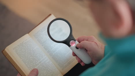 elderly person holding magnifying glass with light, focusing on text in book. hand gently presses button on magnifier, reading small print. soft, blurred background highlighting reading action