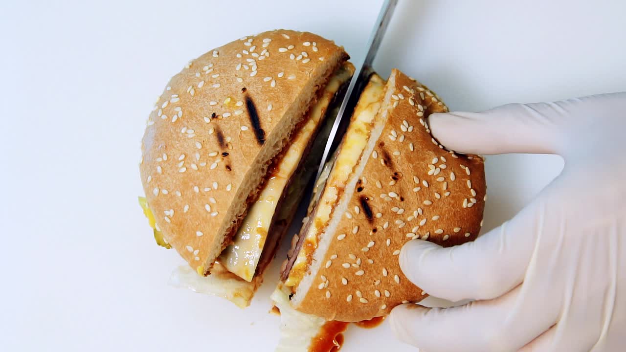 Cutting ready hamburger into halves. Chef with sharp metal knife actively cuts a fresh burger on white background. Top view. Fast food