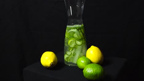gorgeous spinning footage around a glass glossy and reflective carafe filled with water flavored with lime, lemon and cucumber fruit. carafe container with liquid is on a dark black studio background.