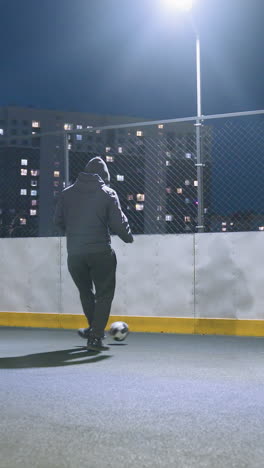 man kicking soccer ball against wall under streetlight on illuminated urban sports field, wall reflects silhouette, showcasing dynamic athletic motion and urban recreational activity at night