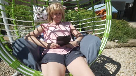 una mujer con pantalones cortos lee una novela digital ebook relajándose en una cómoda hamaca de columpio en un día caluroso y soleado