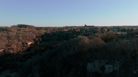 Aerial-shot-with-tracking-shot-of-a-small-village-and-its-church-in-the-middle-of-the-forest-at-sunrise,-Saint-Avit-Ségnieur,-Dordogne