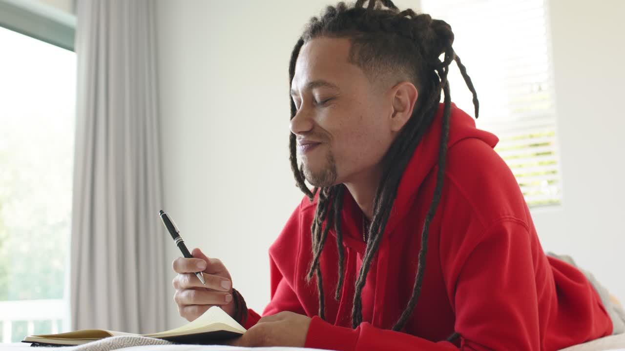 African American man on bed by window tapping pen seeking inspiration writing in notebook smiling