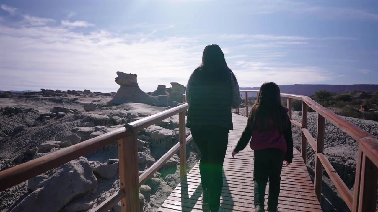 Vista de costas de uma mãe e filha caminhando lado a lado ao longo de um calçadão de madeira através das formações rochosas únicas do Parque Provincial de Ischigualasto, Argentina
