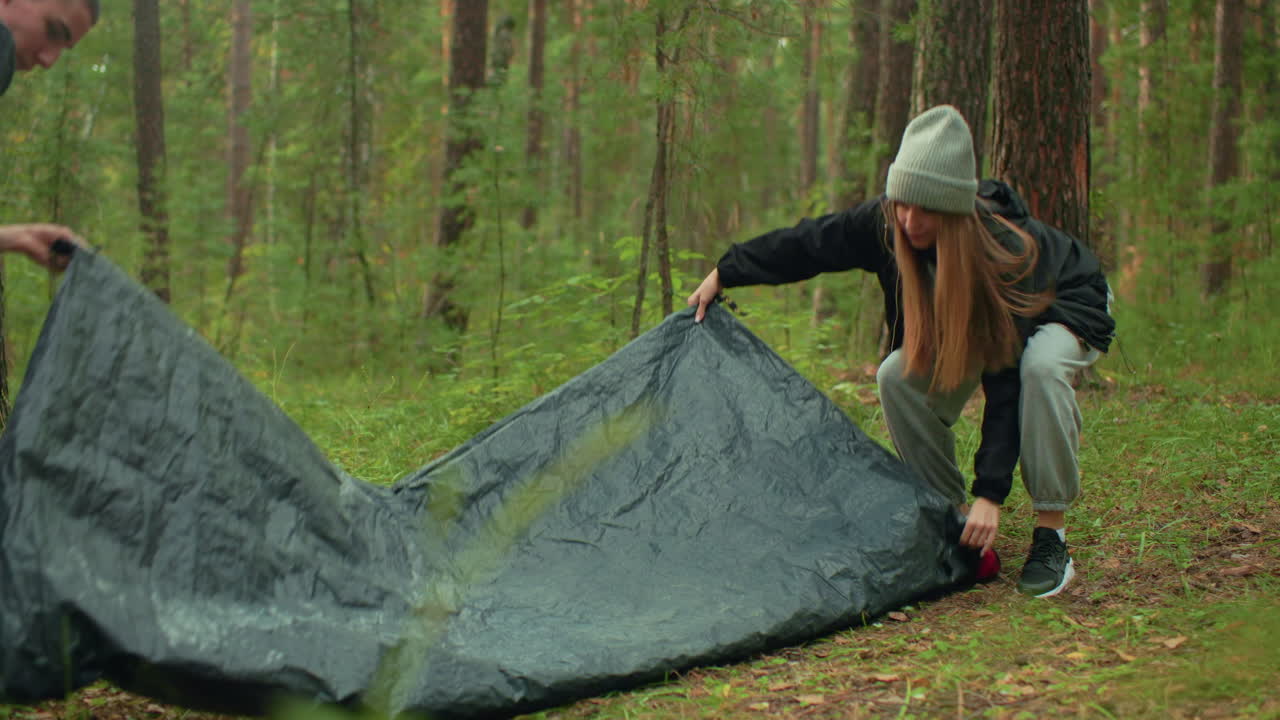 Young lady crouches to pack tent in forest while boyfriend approaches to assist, drawing close as they share gentle kiss surrounded by trees, pine trunks, and soft greenery in serene outdoor setting