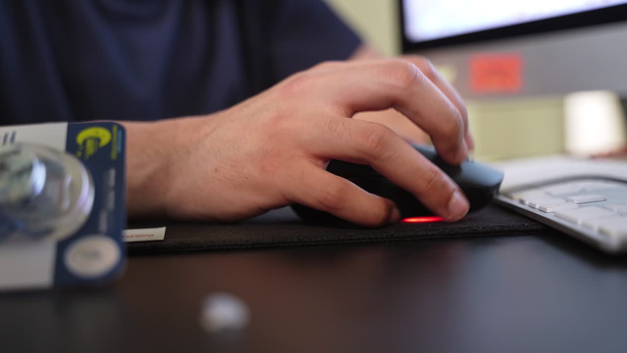 Close-up of a hand operating a computer mouse on an office desk beside a keyboard, with a blurred monitor in the background and soft indoor lighting