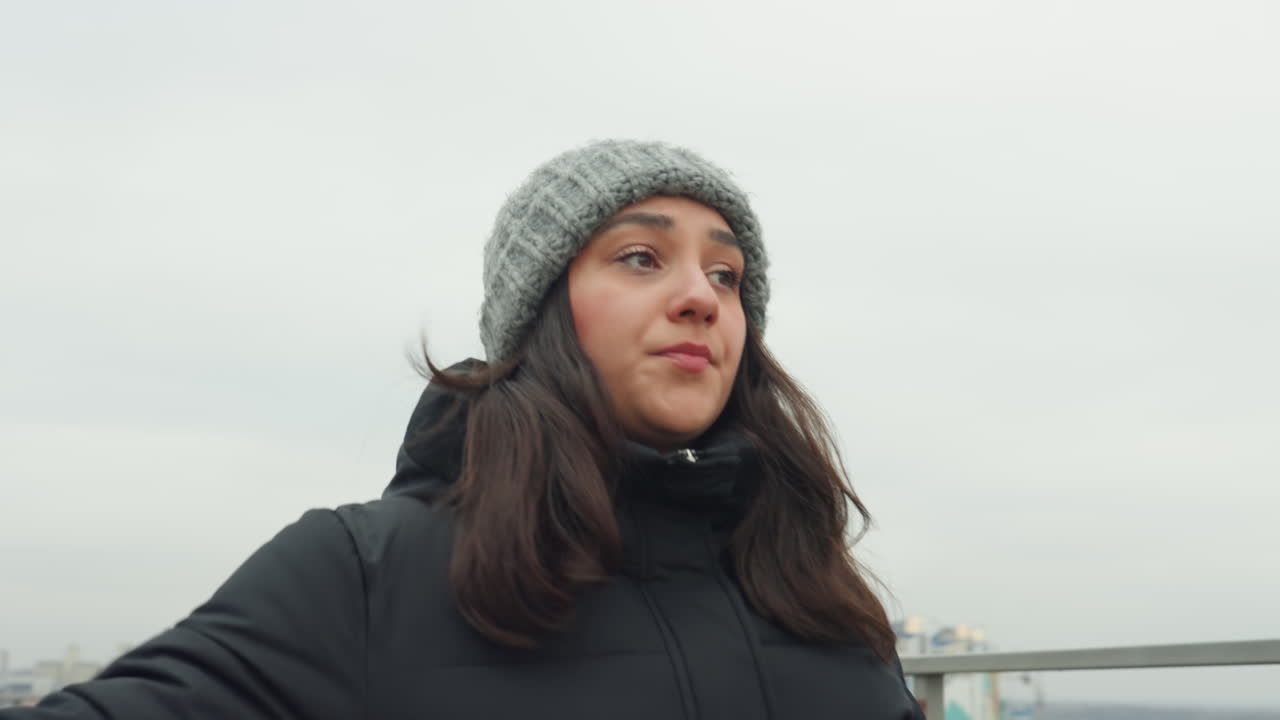 Woman dancing on urban overlook wearing beanie and puffer coat, playful arm swings and steady steps along path, city panorama behind, closeup expressions alternating with wide shots, rhythmic practice