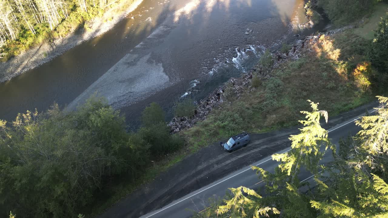 Aerial view of a winding forest road beside a riverbank with morning shadows and natural greenery