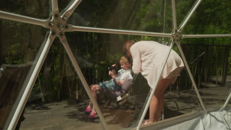 girl and mother look at photographs taken on camera on balcony with glass fence of house surrounded by dense forest. photos for memory and spending time with family