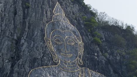 buddha's face engraved with gold into the hill in khao chi chan, thailand. cinematic footage of buddha mountain in pattaya at dawn. pan forward to buddha's golden eyes
