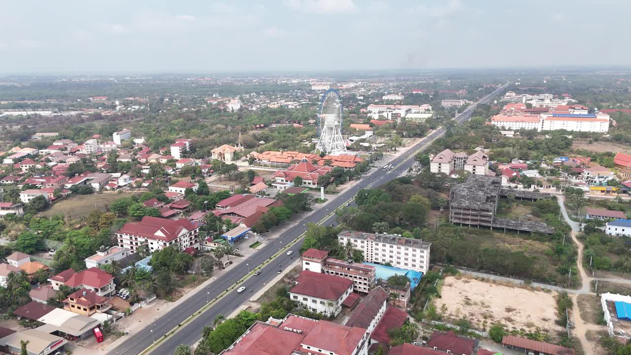 Traditional Cambodian-style roofs and modern multi-story buildings. There are also many trees and areas of greenery throughout the urban landscape. In the background, a tall structure, a Ferris wheel