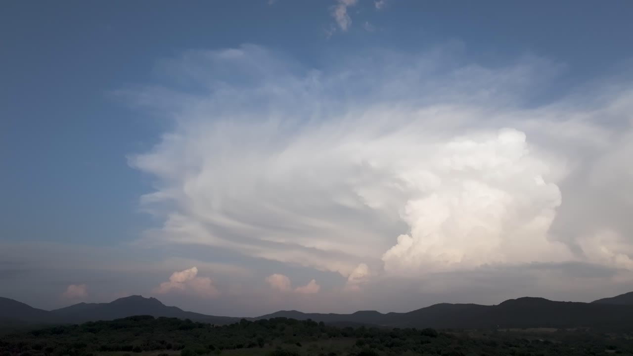 Spectacular lateral drone filming of a large Cumulonimbus cloud created behind a line of mountains and with a blue sky background, it evolves and we appreciate its great width,